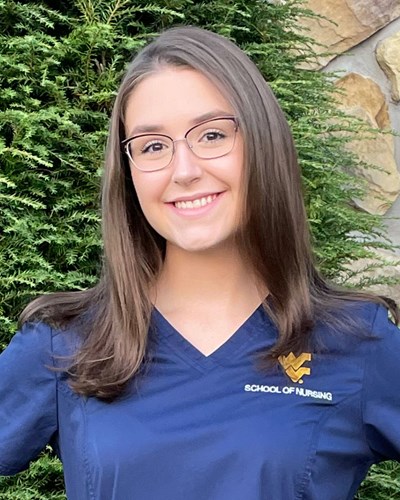 Whitney smiles for the camera while wearing blue WVU School of Nursing scrubs and standing in front of an evergreen shrub and a stone wall.