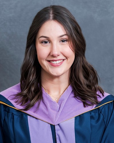 Kayla smiles and poses for a portrait while wearing a blue graduation gown trimmed in purple.