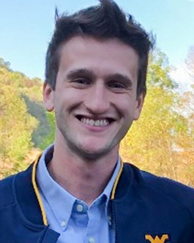 Colton smiles for the camera, wearing a blue WVU jacket and light blue button-down shirt. He is standing in seemingly rural area with a couple of buildings behind him, which are in front of a wooded hillside.