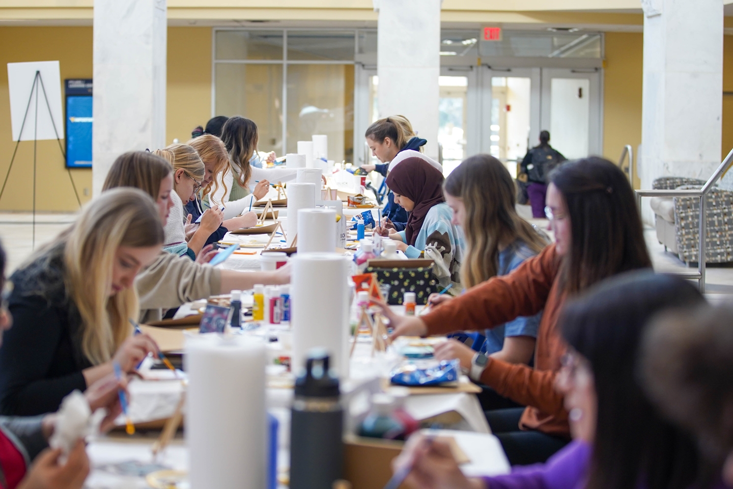A group of people gathered around a long table, painting on small canvases with various art supplies. The setting is a spacious indoor area with large windows and marble columns.
