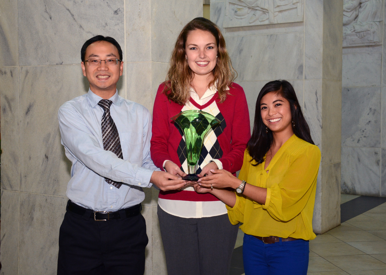 A group of three people showcasing a student award in front of the Health Sciences Center Pylons.
