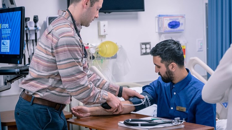 A student, standing in the STEPS Simulation Lab, practices using a blood pressure cuff on a seated student.