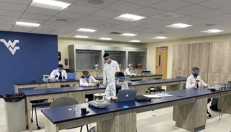 A laboratory classroom is shown with six School of Pharmacy students spread out amongst long desks and using the lab’s various pieces of equipment. The students are all wearing lab coats, face masks, and face shields.