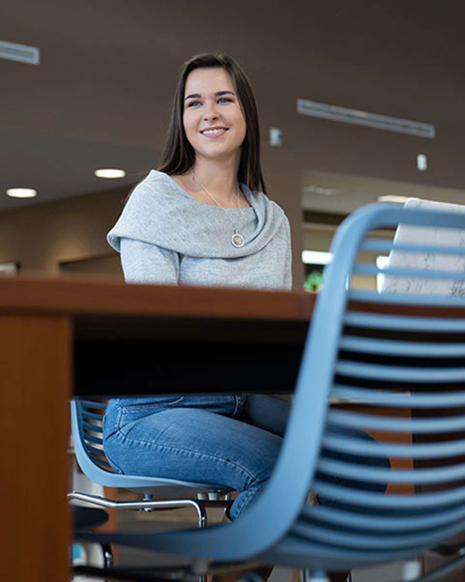 Student seated at a table in the library with her computer smiling while looking out the window.