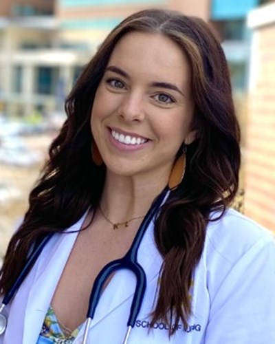 Kasey is seen in a white coat with stethoscope around her neck. She is standing on a set of stairs in between walls, as J.W. Ruby Memorial Hopsital can be seen in the background.