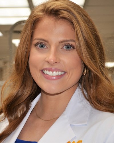 Kaci looks towards the camera while smiling. She is wearing a white WVU lab coat and is posing in a dental facility at the Health Sciences Center.
