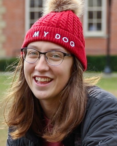 Miranda—wearing a red beanie with “My Dog” stitched in white on the front fold—smiles as she kneels on the ground inside Woodburn Circle.