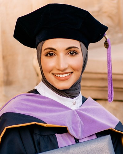 Khadeeja is seen smiling, wearing a purple-highlighted doctoral cap and gown and holding her degree. She is seated on some stairs outside a classroom building.