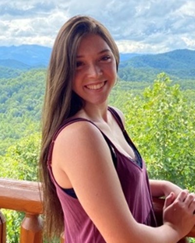 Haley smiles as she turns to look over her shoulder at the camera. Her hand is resting on a wooden railing, and behind her are tree-covered hills and a cloudy sky.