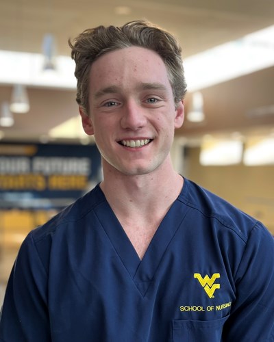 Liam, wearing blue School of Nursing scrubs, smiles as he stands in front of the Pylons of the Health Science Center lobby in Morgantown.