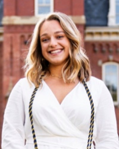 Maggie smiles while standing in the middle of Woodburn Circle, wearing a white dress with honors cords draped around her neck.