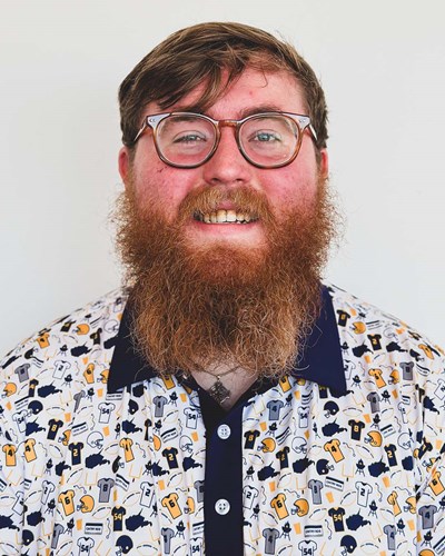 Brandon smiles in a portrait photo. He is wearing glasses, and a buttoned-down shirt that has a busy paattern of Mountaineer football jerseys, helmets, and the shape of the state of West Virginia on it.