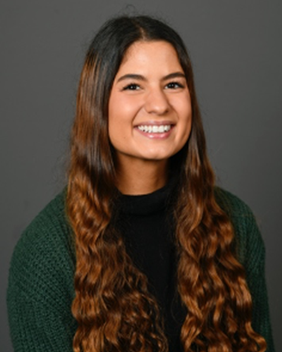 Miranda smiles for the camera. She is posing in front of a dark gray background, and is wearing a green knit sweater.