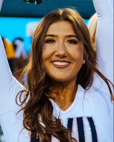 Madison is shown standing on the sidelines of Mountaineer field wearing a cheerleading outfit, with her arms raised and gold and blue pompoms in her hands.
